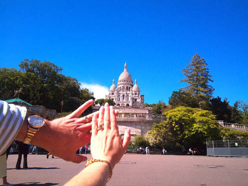 A couple's hands holding up a mirror image of the Sacre-Cœur Basilica in Paris under a bright blue sky, with one person wearing a gold watch and the other a gold bracelet.