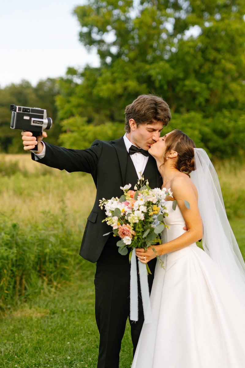 A bride in a white wedding gown and veil and a groom in a black tuxedo kiss while the groom takes a vintage-style film selfie outdoors in a grassy field with trees in the background.
