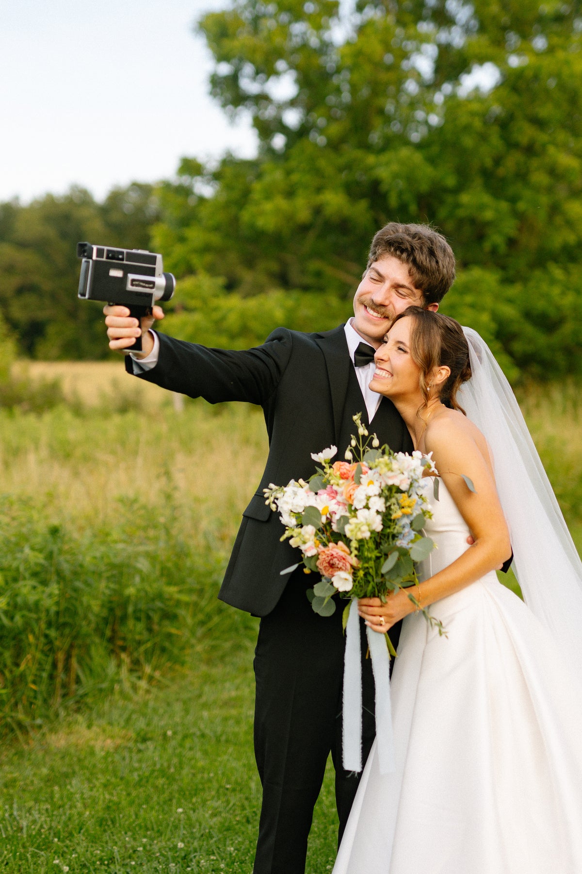 A bride in a white wedding gown and veil taking a vintage-style film selfie with a groom in a black tuxedo jacket outdoors in a grassy field with trees in the background.