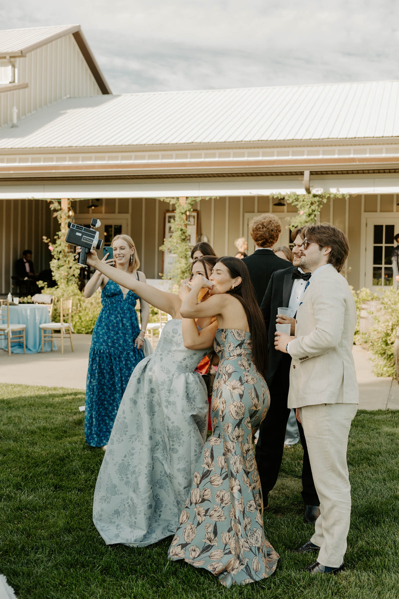 A group of wedding guests taking a selfie outdoors on a lawn in front of a white-roofed building. The central focus is on a woman in a light blue lace gown holding a phone, surrounded by others in formalwear, including a man in a cream suit and a woman in a blue patterned dress.